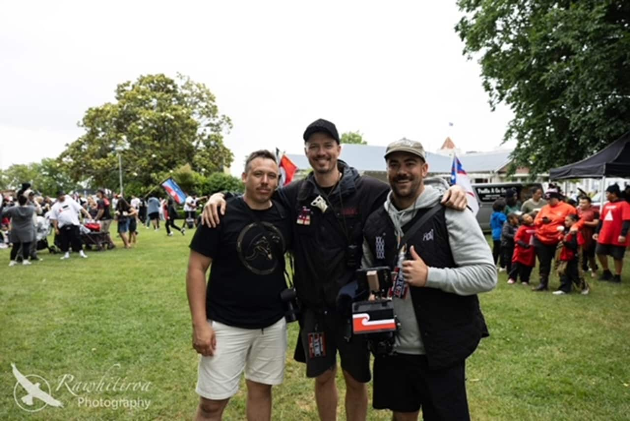 Three men smile for the camera, with the man in the middle resting his arms on the shoulders of the two men standing beside him.