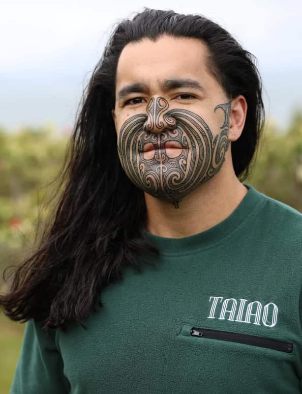 A man with shoulder-length black hair and face tattoos wearing a green T-shirt.