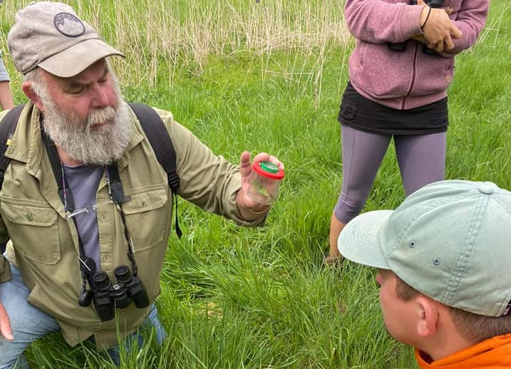 A man in green vest and hat and with black binoculars around his neck kneels and holds up a small pot in which there is an insect. A person in a cap looks on