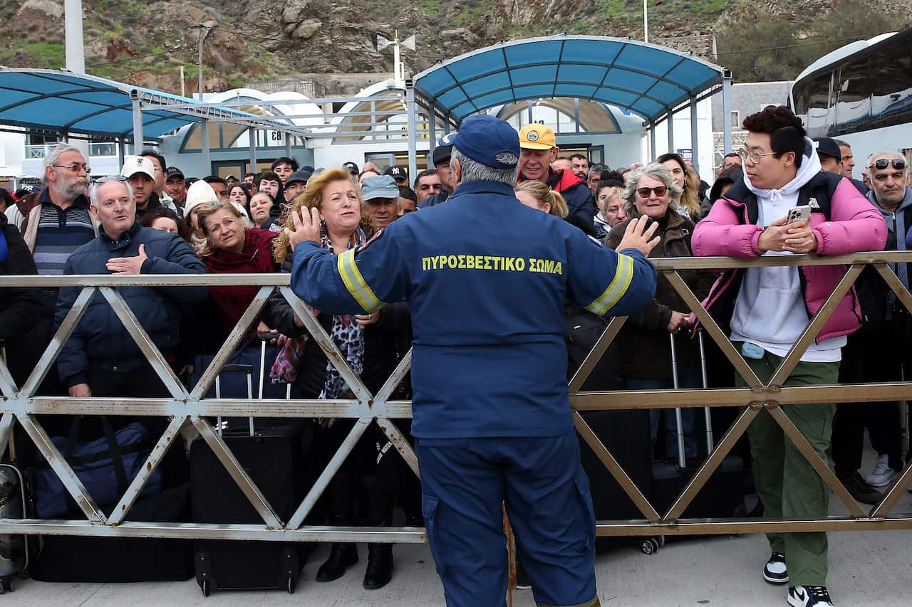 A ferry worker in blue overalls with his back towards the camera gestures with his hands, urging a group of people standing on the opposite side of a wooden barricade to wait for their turn.